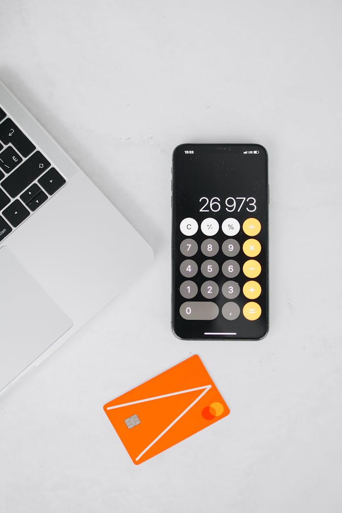 A flat lay of a laptop, smartphone with calculator app, and orange card on a white surface.